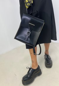 Black leather backpack with a structured design, featuring a buckle closure, yellow stitching, and a single adjustable strap. Black platform shoes underneath.