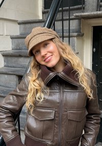 Casaco bomber de couro castanho com gola canelada castanha e bolsos, combinando com um boné castanho de tweed, em frente a degraus de pedra.