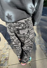 Persoană care merge pe o stradă pavată, îmbrăcată în pantaloni negri cu modele grafice albe și un hanorac negru cu desene de stele.