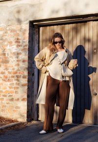 Beige Trenchcoat über einem cremefarbenen Strickoberteil, braunen, weiten Hosen und weißen Sneakern. Die Accessoires umfassen eine kleine, fransige Tasche und Sonnenbrillen.