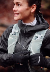 Veste imperméable noire avec des accents réfléchissants, associée à un gilet d'hydratation vert clair. Les gants sont noirs avec un détail de logo blanc.
