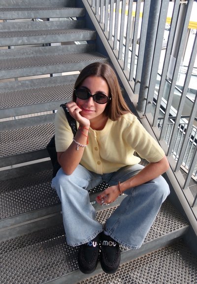 Cárdigan amarillo de punto con mangas cortas, jeans de pierna ancha azul claro y zapatos negros sin cordones, sentado en escalones de metal. Gafas de sol negras.