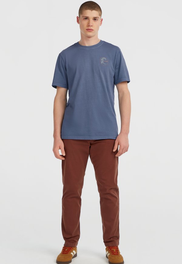 Blå bomulds T-shirt med rund halsudskæring og korte ærmer, der har et lille logo på brystet. Bæret med brune bukser og brune sneakers.