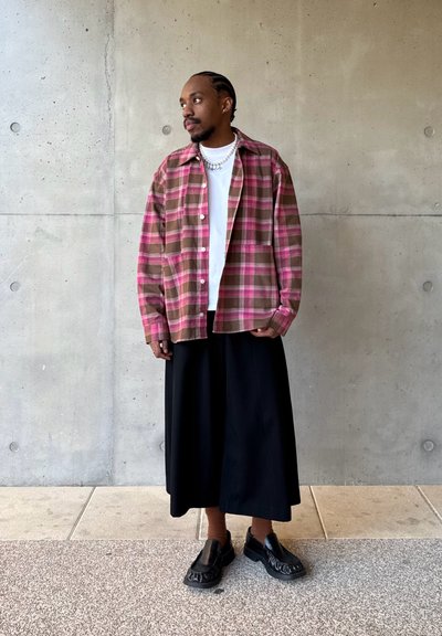 Hombre con trenzas que lleva una chaqueta de cuadros rosa, camisa blanca, falda larga negra, calcetines marrones y zapatos negros, de pie contra una pared de hormigón gris.