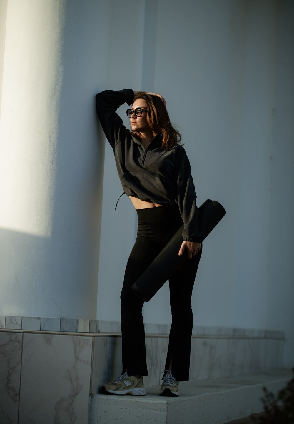 Black cropped jacket, high-waisted black leggings, and light-coloured athletic shoes. Holding a rolled yoga mat. Solid background and columns.