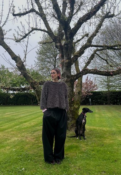 Suéter gris y difuso con mangas amplias, pantalones negros de gran tamaño, de pie junto a un perro negro, bajo un árbol en un entorno exterior con hierba.