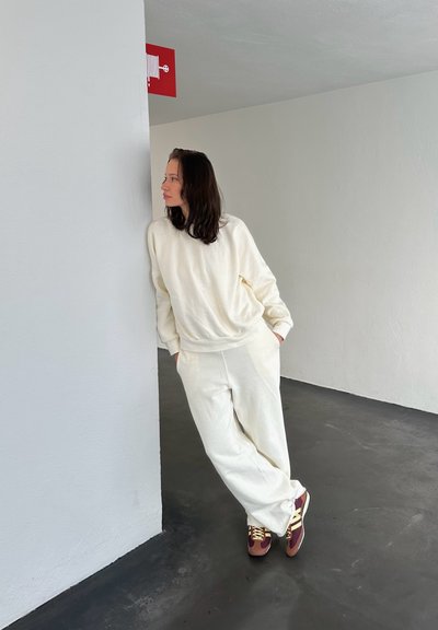 Sudadera de tela suave color crema y pantalones a juego, de corte relajado. Zapatillas marrones y burdeos con acentos dorados, frente a una pared gris.