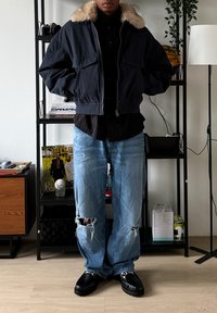 Veste bomber marine avec col en fausse fourrure, chemise noire à rayures, jean bleu clair délavé et chaussures noires. Coutures apparentes sur le jean.