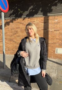 Femme blonde en manteau matelassé noir et pull gris, debout sur un trottoir ensoleillé près d'un mur en briques et d'un panneau d'interdiction de stationner, tenant un sac noir.
