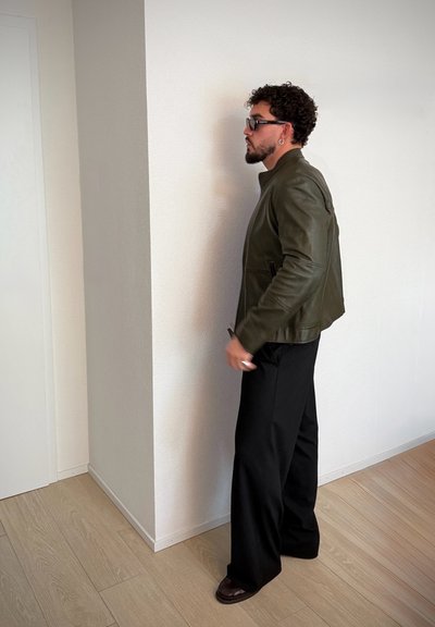Chaqueta de cuero verde oliva con cuello alto, combinada con pantalones anchos negros. La escena presenta un suelo de madera clara contra una pared blanca.
