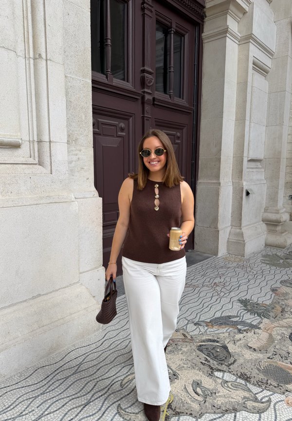 Femme portant un haut sans manches marron et un pantalon blanc, tenant un verre et un sac à main, debout sur un pavement en pierre à motifs près d'une grande porte en bois sombre.