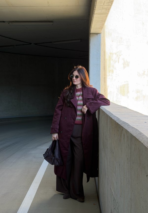 Mujer con un abrigo largo burdeos, suéter a rayas, gafas de sol y sosteniendo un bolso, apoyada contra una pared de hormigón en un garaje.