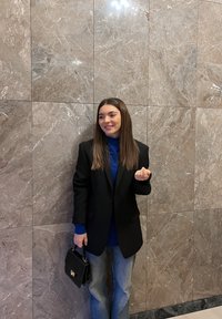 Žena má na sebe čierny blejzer cez modrý rolák, v kombinácii so svetlomodrými džínsami. Drží čiernu kabelku. Na pozadí je mramorová stena.