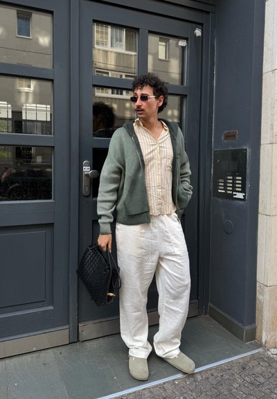 Persona que lleva un cárdigan verde oliva claro sobre una camisa de rayas verticales crema, pantalones blancos holgados y zapatos grises sin cordones, sosteniendo un bolso negro.
