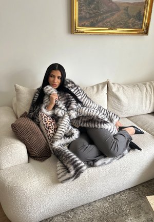 Mujer de cabello largo y oscuro, vestida con un abrigo de piel a rayas y pantalones grises, recostada en un sofá crema con cojines estampados, bajo una pintura de paisaje.