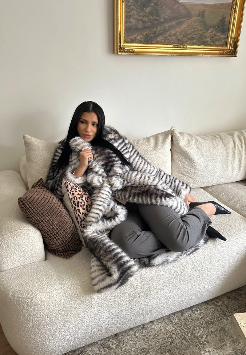 Femme aux longs cheveux foncés portant un manteau en fourrure rayé et un pantalon gris, allongée sur un canapé crème avec des coussins à motifs sous une peinture de paysage.