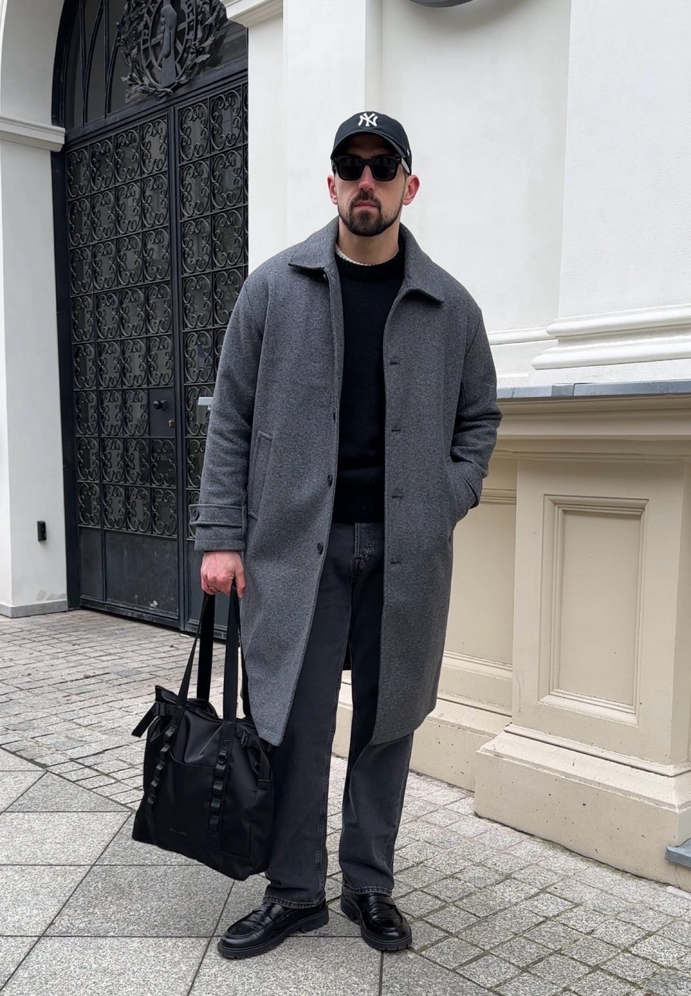 Cappotto di lana grigio su un maglione nero, jeans neri e scarpe di pelle nere. Porta una borsa nera con dettagli a nastro. Indossa occhiali da sole e cappello.