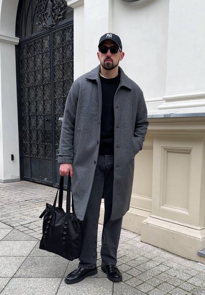 Abrigo de lana gris sobre un suéter negro, jeans negros y zapatos de cuero negros. Sosteniendo una bolsa negra con detalles de cinta. Lleva gafas de sol y gorra.