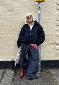 Denimová bunda, bílý tričko, červený šátek, oversized denimové kalhoty a červené kotníkové boty. Krémová čepice a doplňky v ruce; stojí vedle sloupu.