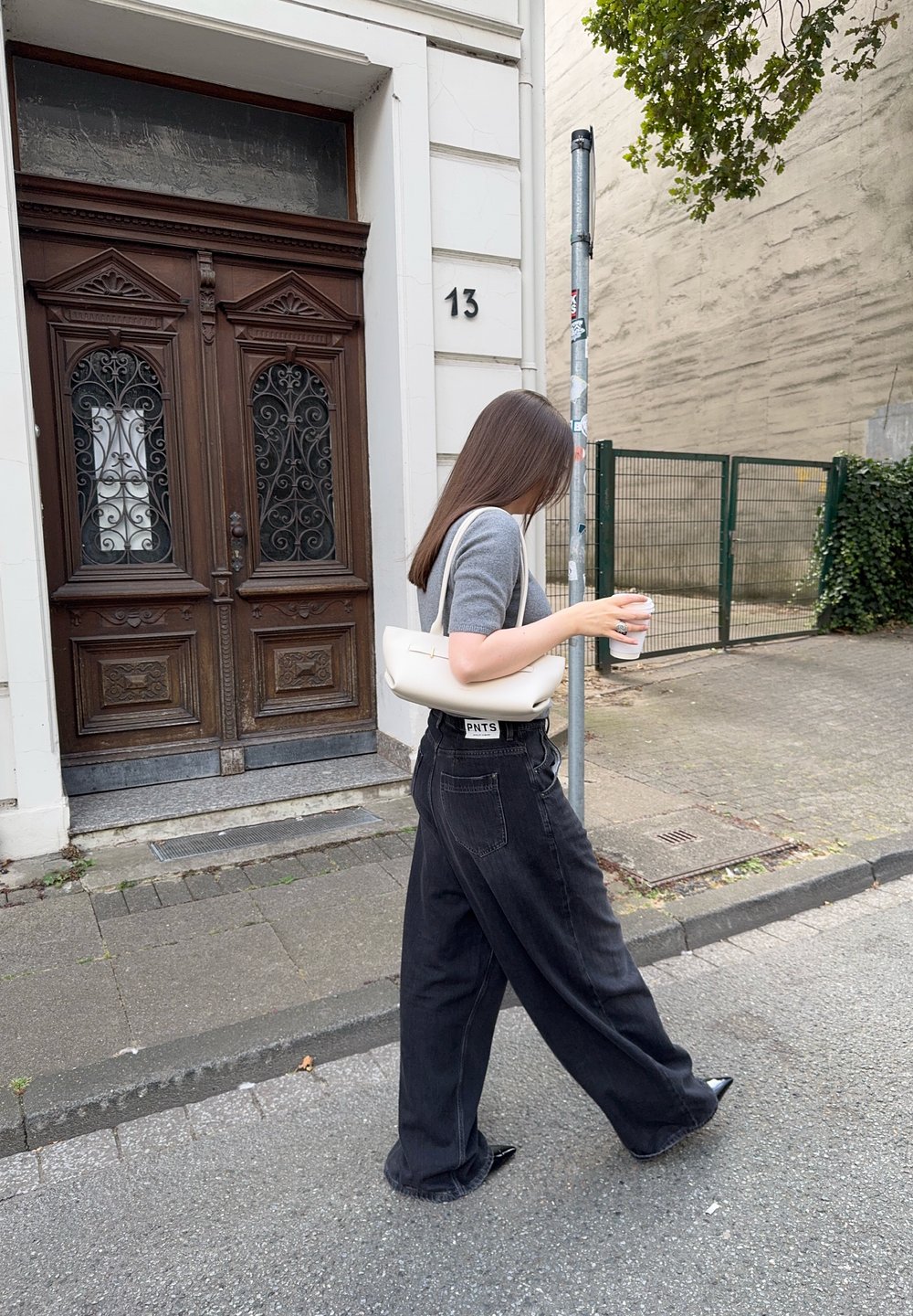 Una persona camina por una acera, vistiendo una camiseta gris de manga corta y pantalones negros holgados, llevando una taza de café y un bolso blanco.