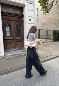 Una persona camina por una acera, vistiendo una camiseta gris de manga corta y pantalones negros holgados, llevando una taza de café y un bolso blanco.