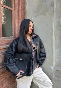 Černá kožená bunda s oversized rukávy, přesažená přes kostkovanou košili. Drží černou koženou kabelku se stříbrným akcentem.