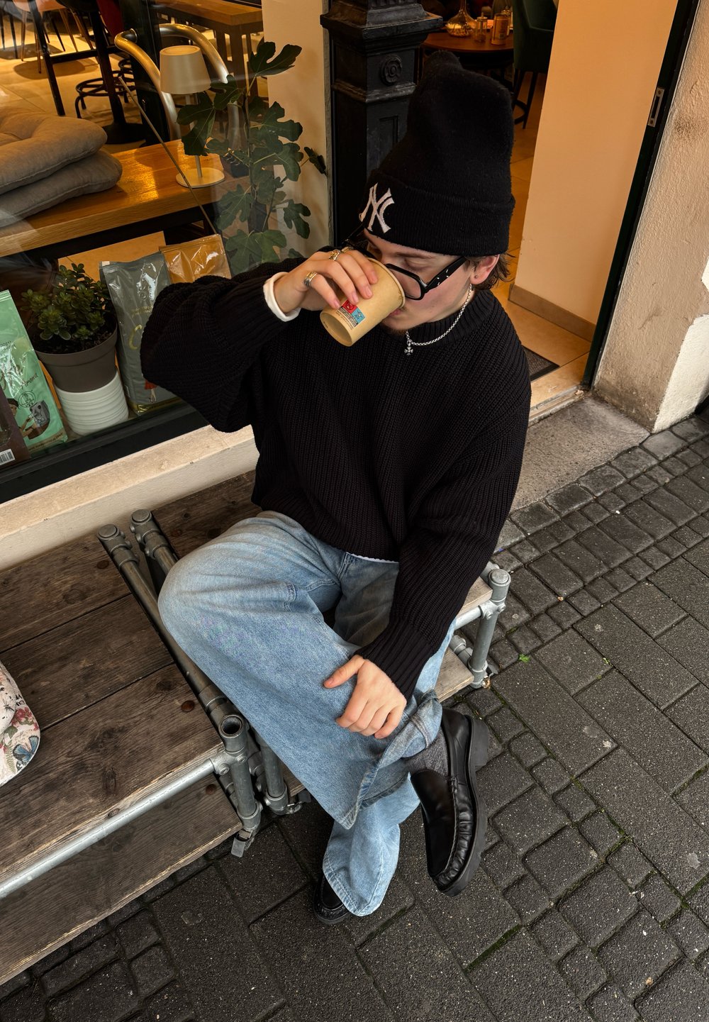 Oseba v črnem oversize pletenem puloverju, ohlapnih svetlo modrih jeansih in črnih škornjih, drži kartonsko skodelico in sedi na leseni platformi.