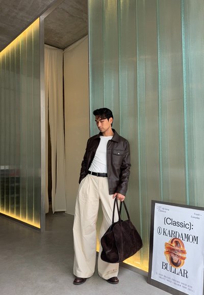 Hombre con camisa blanca, chaqueta de cuero marrón y pantalones beige anchos sosteniendo una gran bolsa de ante marrón oscuro, de pie junto a una pared de cristal y un cartel de pastelería.