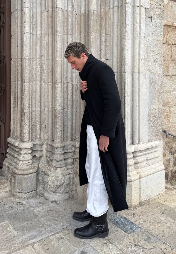 Persona con cappotto lungo nero, pantaloni bianchi e stivali neri in piedi accanto a una colonna di pietra dettagliata su un terreno lastricato, mentre guarda verso il basso.