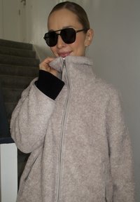 Veste en polaire duveteuse rose clair avec col montant et fermeture éclair, dotée de poignets en tissu foncé et de fermetures éclair latérales, portée avec des lunettes de soleil noires.