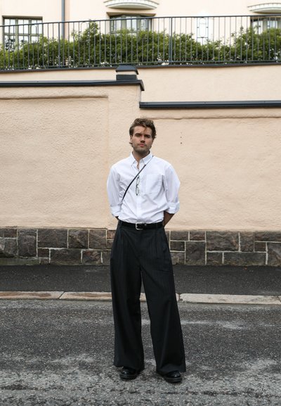 Camisa blanca de botones, pantalones anchos de rayas negras, cinturón negro, gafas colgando de un cordón, de pie en una calle texturizada.