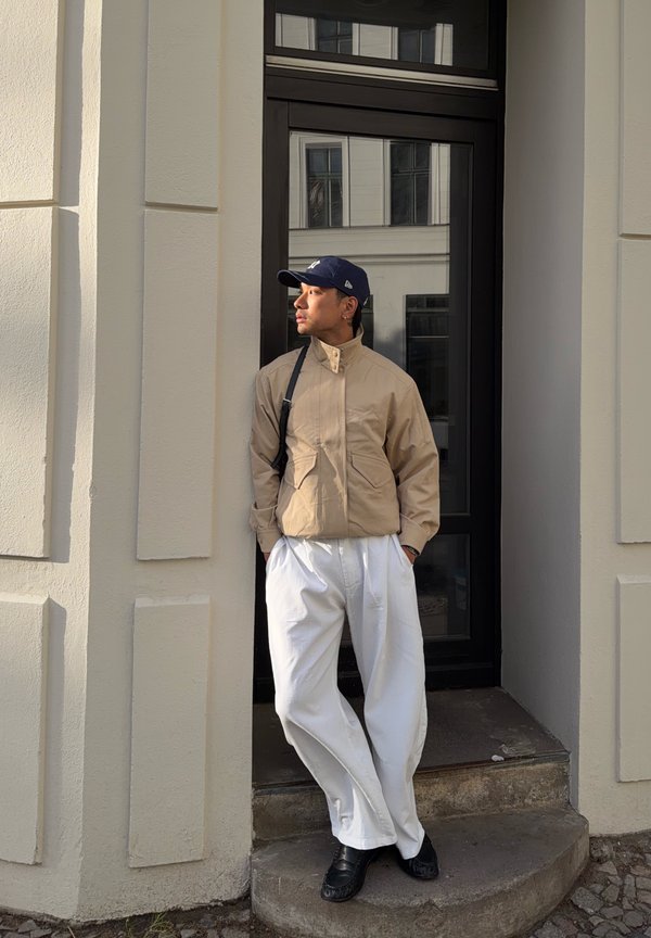Homme portant une veste beige, un pantalon blanc, des chaussures noires et une casquette marine, se tenant les mains dans les poches près d'une porte noire dans un bâtiment clair.