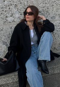 Γυναίκα που φοράει μαύρο παλτό, γκρι πουλόβερ με φερμουάρ, λευκό τοπ, μπλε τζιν, μαύρες μπότες και μεγάλα μαύρα γυαλιά ηλίου, καθισμένη δίπλα σε μια υφή πέτρινος τοίχος.