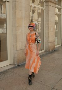 Oranje jurk met wit gemarmerd patroon, mouwloos, met een keyhole halslijn. Gedeeld met zwarte platformlaarzen en een versierde handtas.