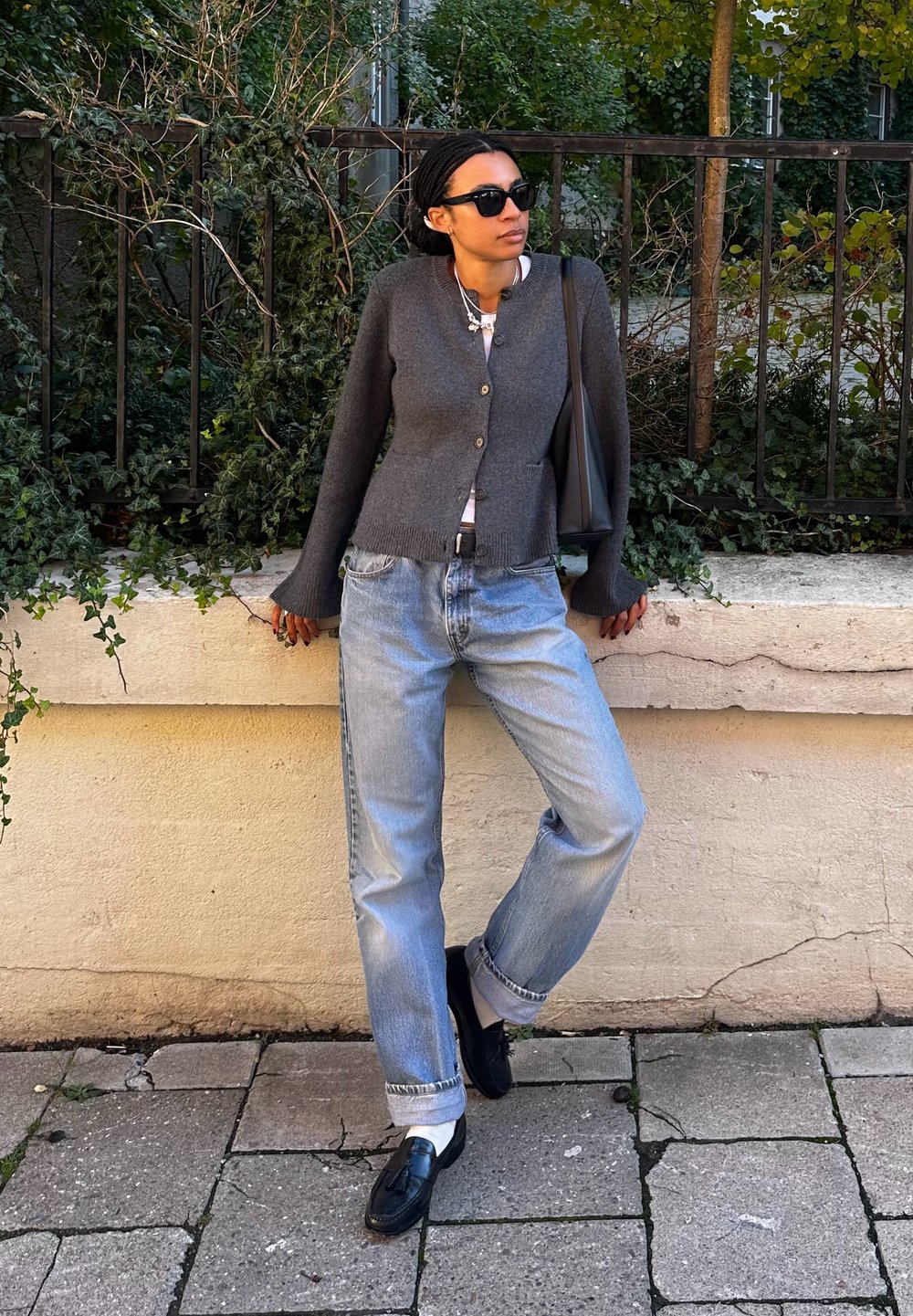 Γκρι ζακέτα με κουμπιά, λευκό πουκάμισο, χαλαρά ανοιχτό μπλε τζιν και μαύρες loafers. Είναι ορατά επίσης γυαλιά ηλίου και μια μαύρη τσάντα.