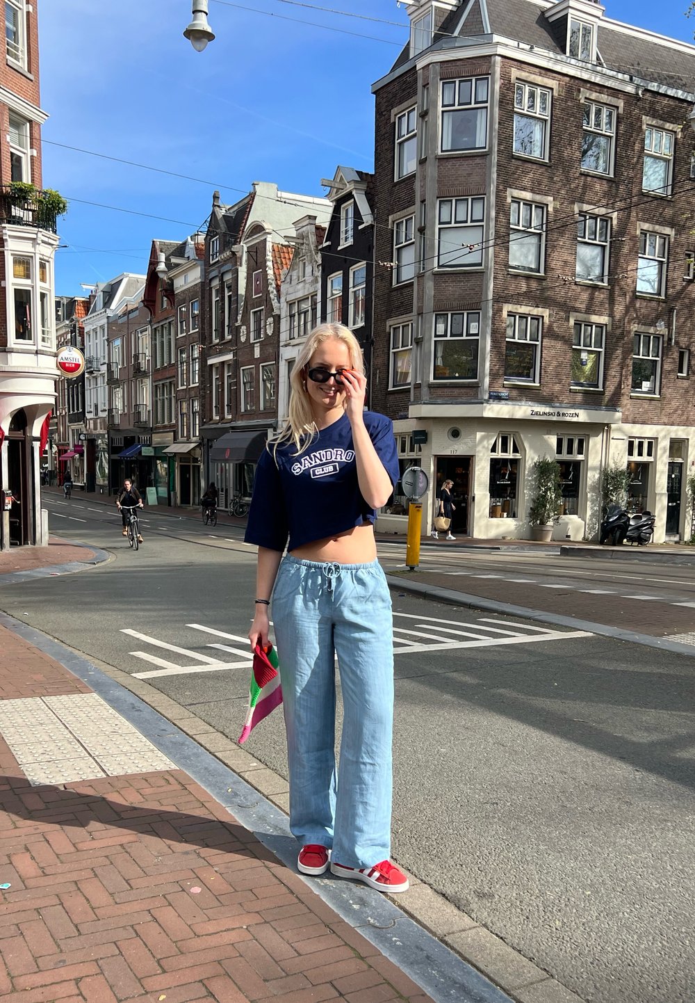 Avskuren marinblå topp med "SANDRO"-logotyp, ljusblå vida byxor, röda sneakers, hållande en flerfärgad väska. Urban gatumiljö med byggnader.