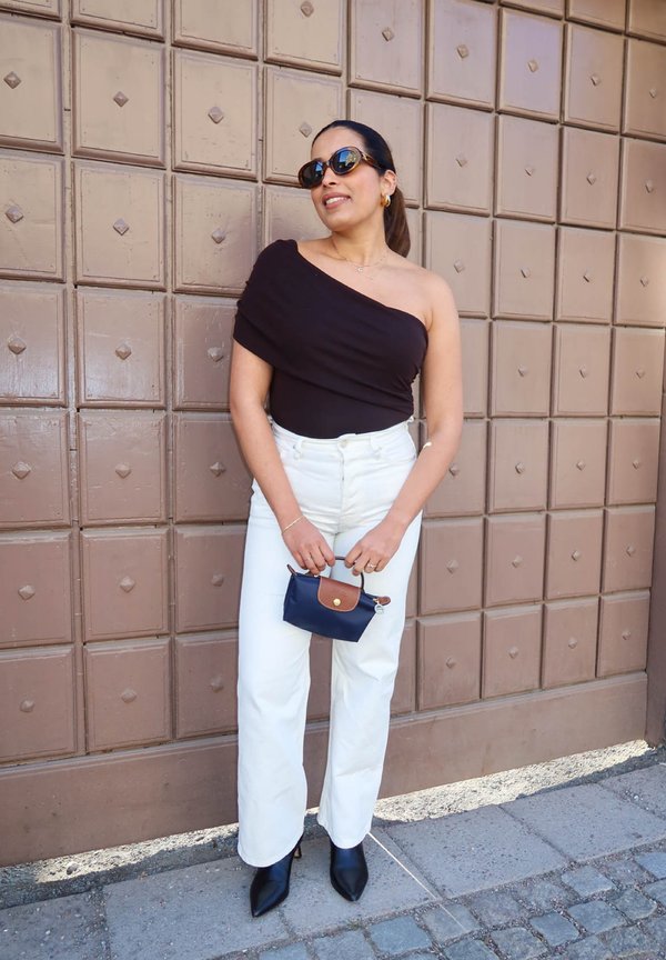 Sieviete melnā viena pleca topā, baltās biksēs, melnās smailītēs zābakos, saulesbrillēs, tur mazu tumši zilu somiņu, stāv pie brūnām durvīm.