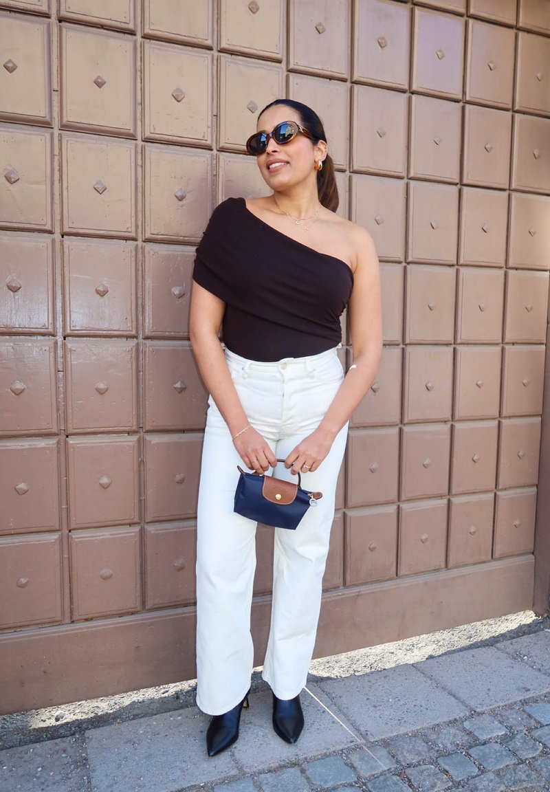 Femme portant un haut noir à une épaule, pantalon blanc, bottines noires pointues, lunettes de soleil, tenant un petit sac à main bleu marine, debout devant une porte marron.