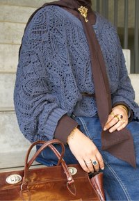 Cardigan bleu en maille ajourée sur une chemise marron, associé à un jean. Sac à main en cuir marron, bijoux en or, bagues et accent foulard.