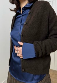 Barna kötött kardigán kék szatén gombos ing felett. A kardigán kerek gombokkal rendelkezik; az ing fényes felületű és hosszított mandzsettás.