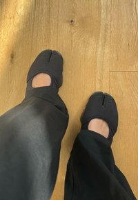 Mustad mustad kingad tekstureeritud kangast, avatud varbaga ja keskelt kulgevate õmblustega. Kanda mustade pükstega puidust põrandal.