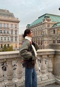 Ung kvinde i grøn jakke og hvidt tørklæde står på en stensal med udsigt over historiske europæiske bygninger på en skyet dag.