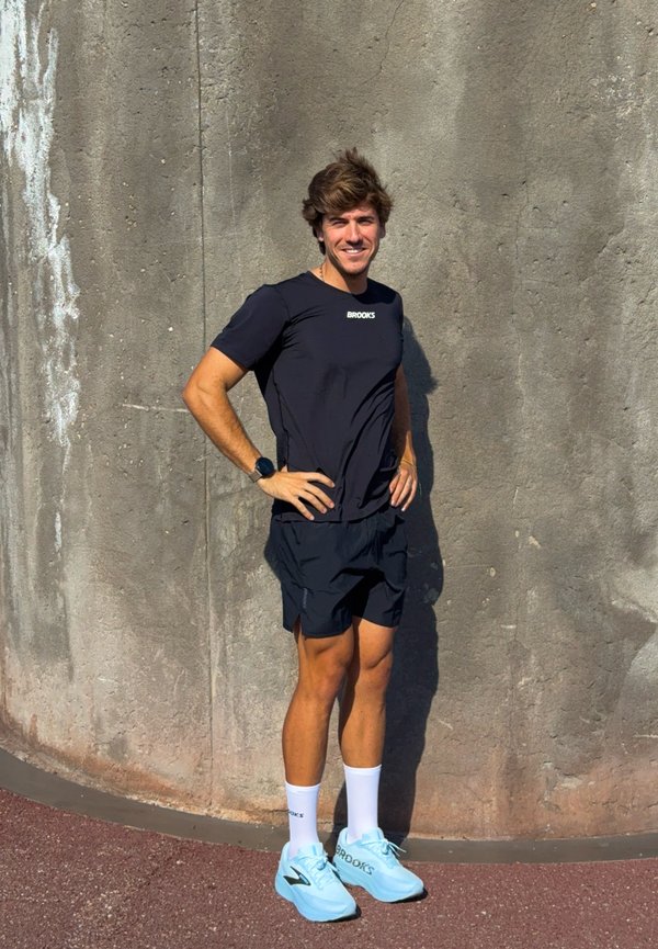 Un hombre sonriente, vestido con ropa negra para correr y calcetines blancos, posa con las manos en las caderas frente a una pared de hormigón. Lleva zapatillas de correr de color azul claro.