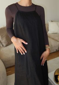 Μαύρο ριγέ φόρεμα με λεπτές τιράντες, φορεμένο πάνω από μια εφαρμοστή σκούρα μπλούζα. Λείο υλικό, ως το γόνατο, με κάθετες γραμμές. Το χέρι ακουμπά στο φόρεμα.