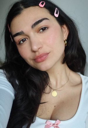 Mujer con cabello largo y oscuro, lleva pinzas de cabello rosas, pendientes de aro dorados, un collar con colgante dorado y una blusa blanca con un lazo rosa.