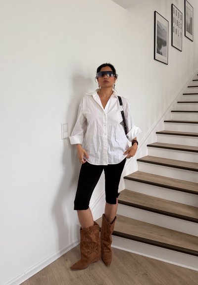 Mujer con camisa blanca de gran tamaño, pantalones negros recortados, botas vaqueras marrones y gafas de sol oscuras, de pie junto a una escalera con cuadros en la pared.