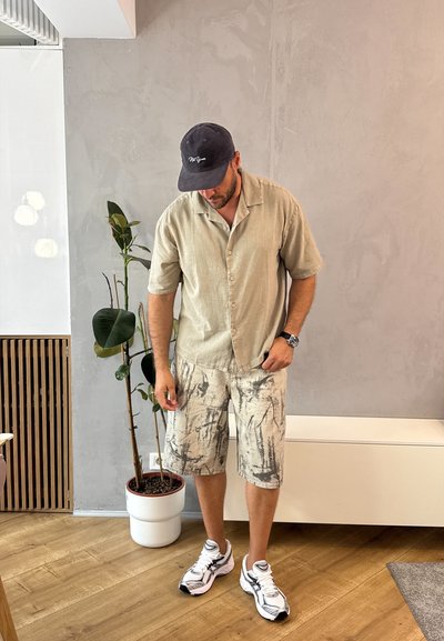 Camisa beige claro de manga corta, pantalones cortos claros con estampado, zapatillas blancas con detalles en negro y una gorra azul marino. Entorno interior con plantas.