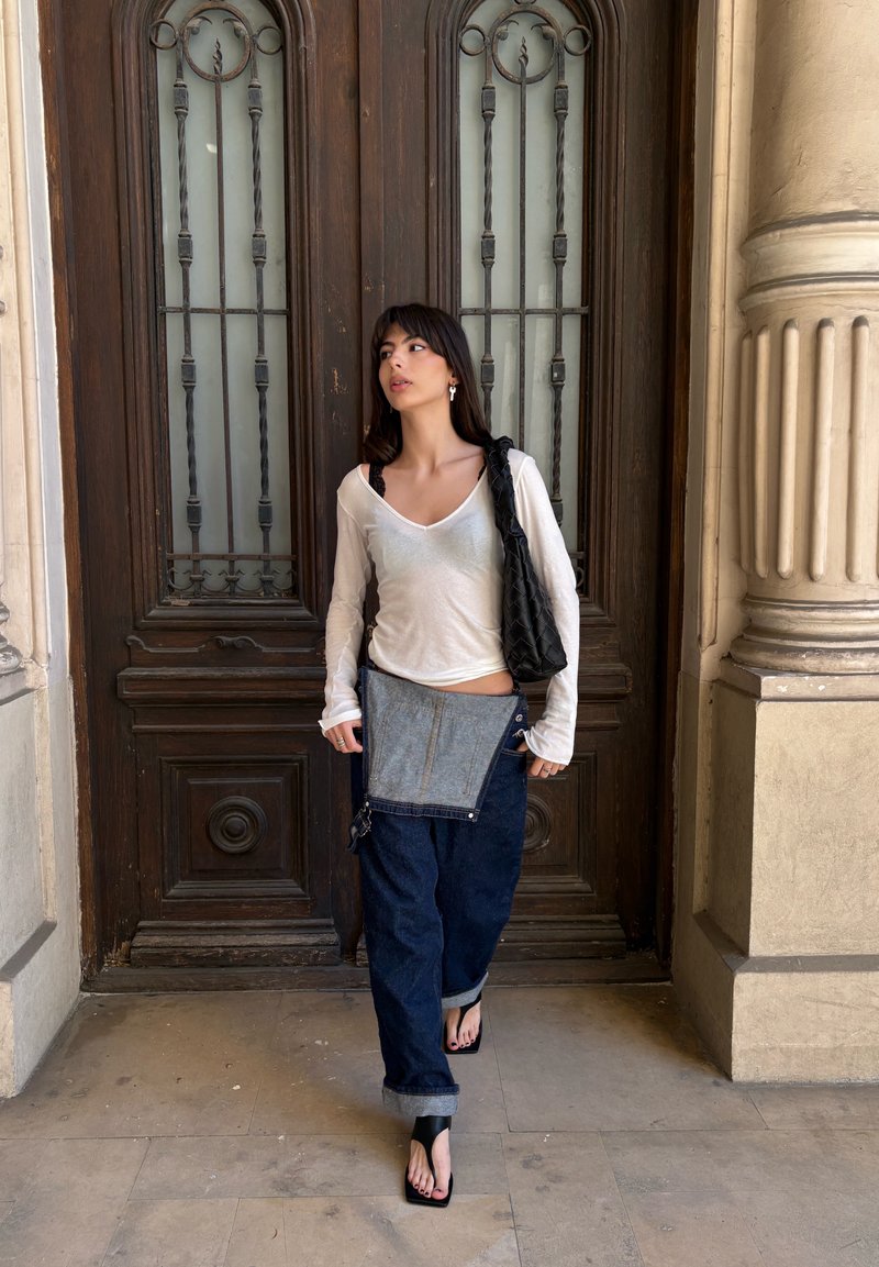 Une femme se tient devant de grandes portes doubles en bois, vêtue d'un haut blanc à manches longues, d'un pantalon marine avec un rabat en denim, de sandales noires et portant un sac noir.