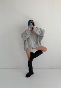 Překročně velká šedá mikina s kapucí, s širokými rukávy a kapsou. Nošena s černými kozačkami po kolena na světlém pozadí.