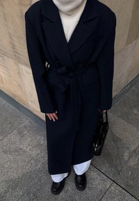Manteau long croisé bleu marine avec une ceinture à nouer, porté sur un pull à col roulé crème, assorti à un pantalon blanc et des bottines noires, tenant un sac noir.